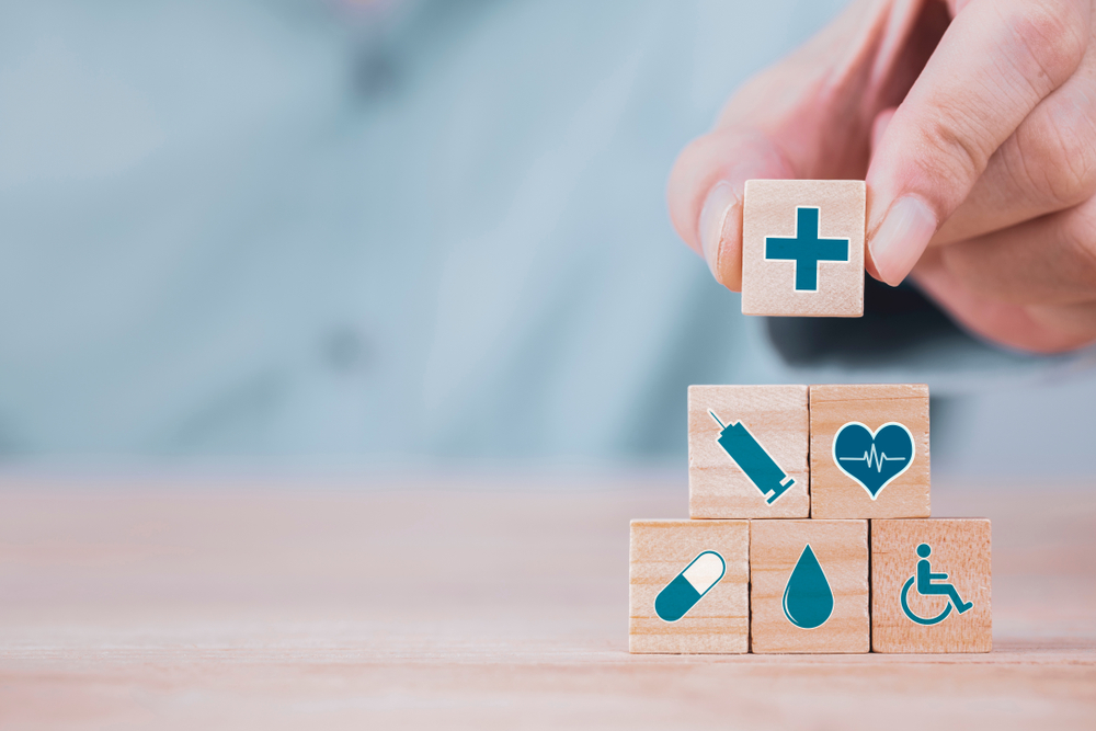 Person adding a wooden building block with a plus sign on it to a pile of blocks with heathcare and medical symbols on them