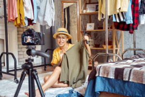 Girl wearing a hat and glasses, sitting at home with a range of clothes she's photographing to sell online